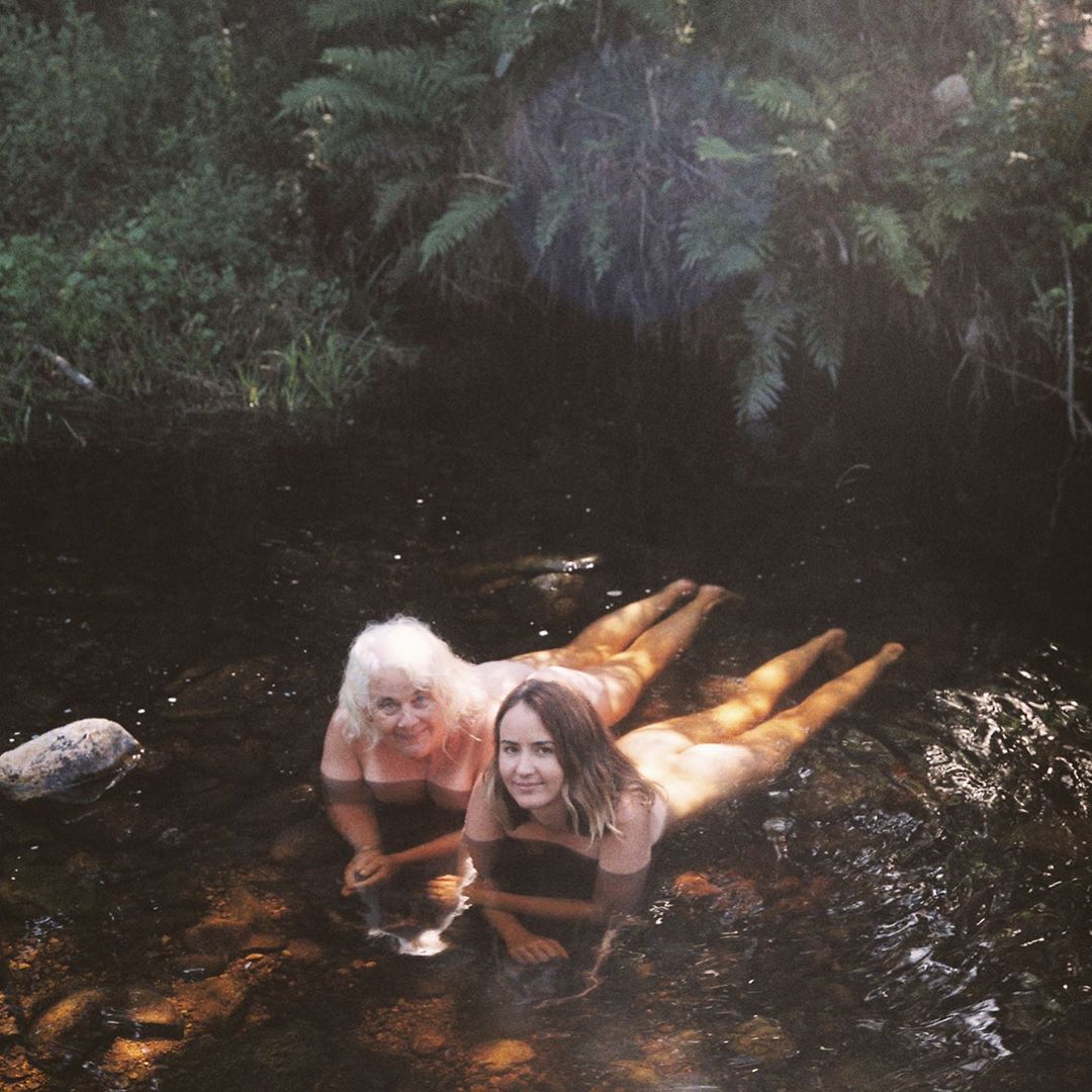 Two women lie in shallow water surrounded by lush green foliage. One woman has white hair, and the other has brown hair, both are smiling.