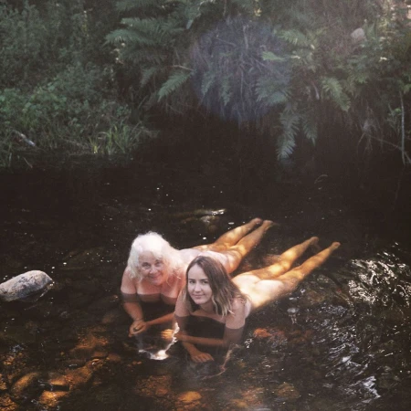 Two women lie in shallow water surrounded by lush green foliage. One woman has white hair, and the other has brown hair, both are smiling.