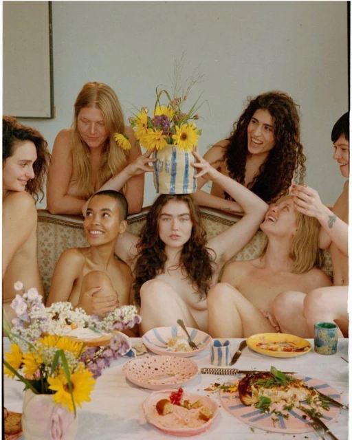 Group of nude adult females gathered around a table with food and flower arrangements in an indoor setting, photographed with natural light.