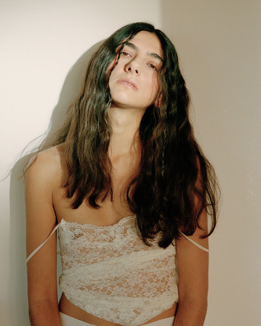 Portrait of a young adult with long brown hair, wearing a lace top, posing in front of a plain white wall with subtle shadow.