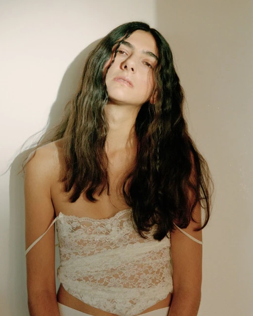 Portrait of a young adult with long brown hair, wearing a lace top, posing in front of a plain white wall with subtle shadow.
