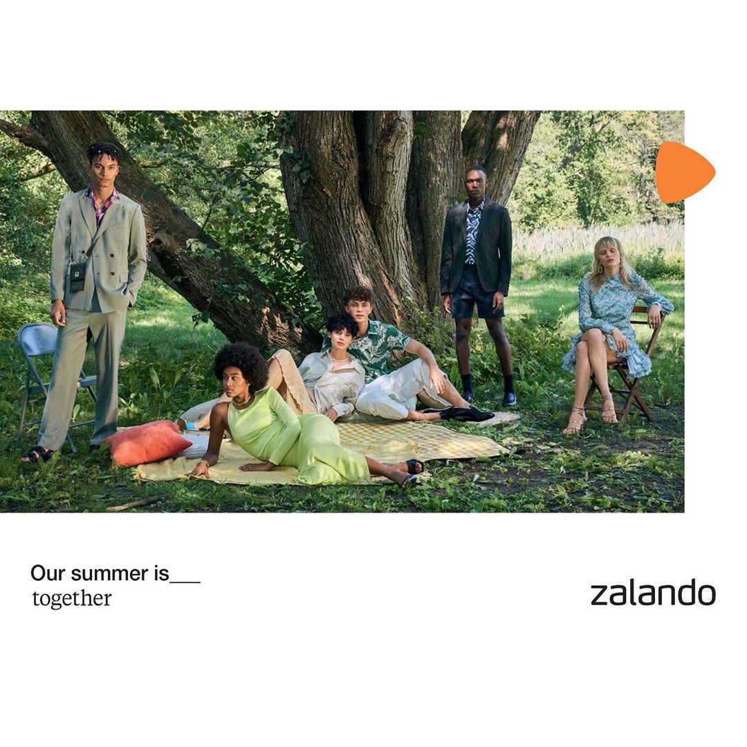 Group of casually dressed young adults relax outdoors in a verdant park, some lounging on a blanket under a large tree in sunny daylight.