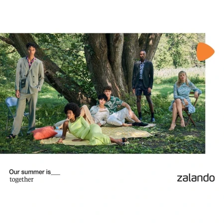 Group of casually dressed young adults relax outdoors in a verdant park, some lounging on a blanket under a large tree in sunny daylight.
