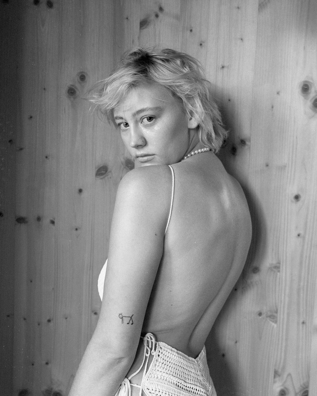 Portrait of a fair-skinned woman with short blonde hair in a white top, looking over her shoulder, with a wood panel background.
