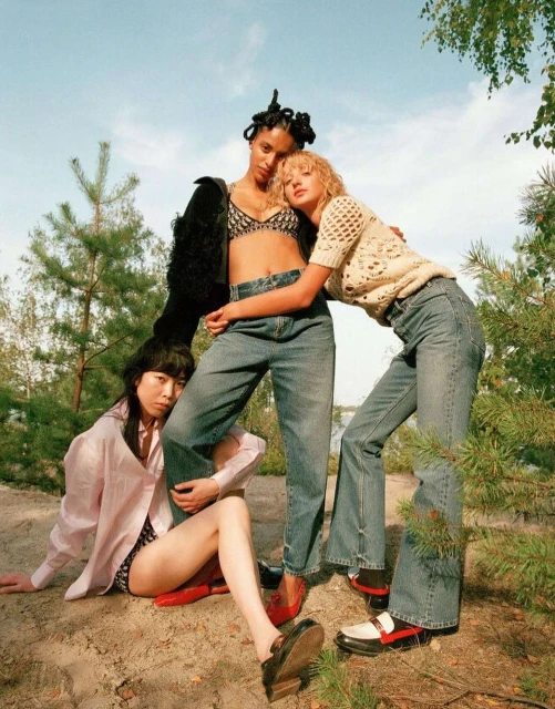 Three women in a field wearing jeans, posing together under a blue sky surrounded by pine trees during daytime in a fashion editorial.