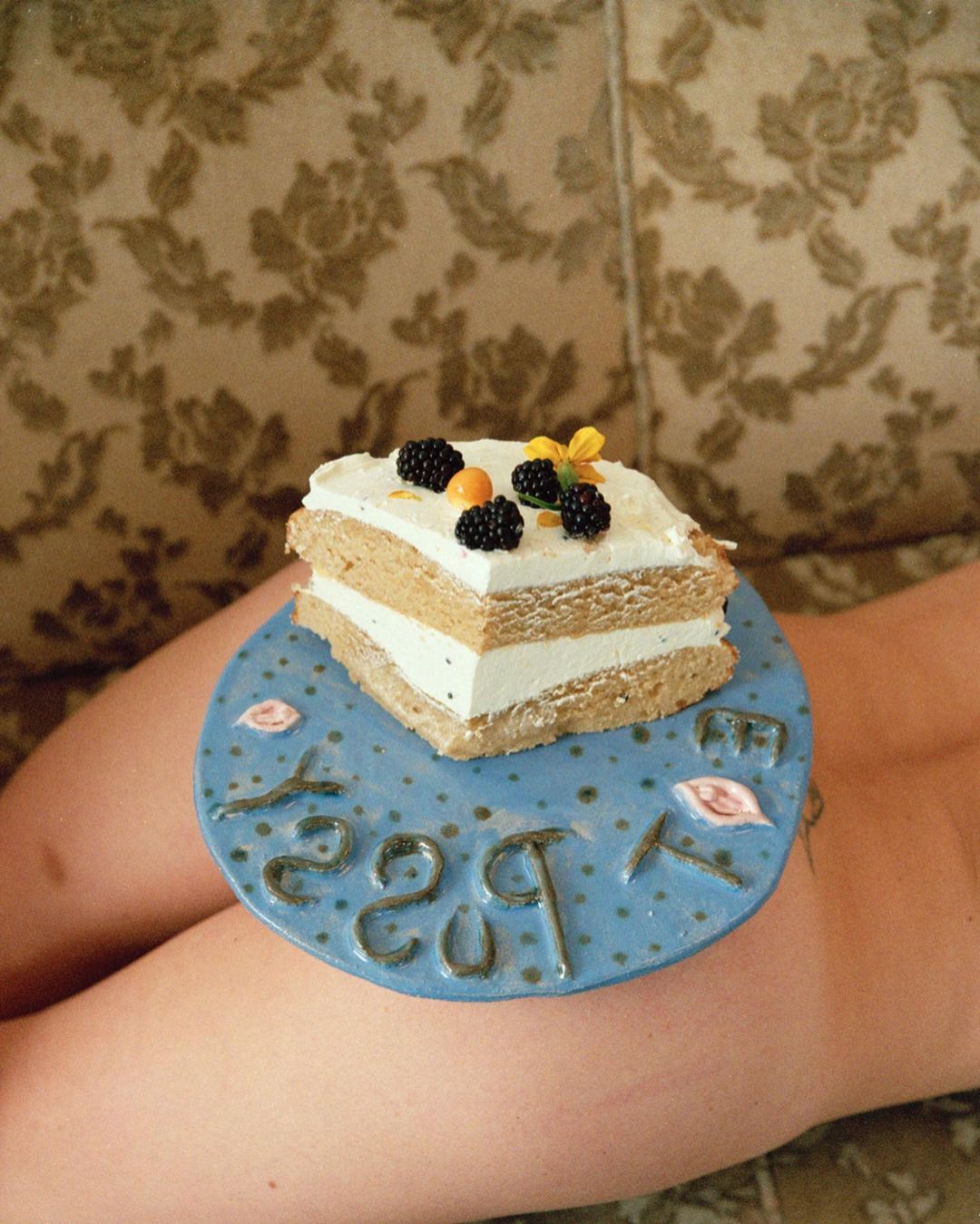 Overhead shot of a layered cake topped with blackberries and a yellow flower on a blue ceramic plate resting on a person's lap.