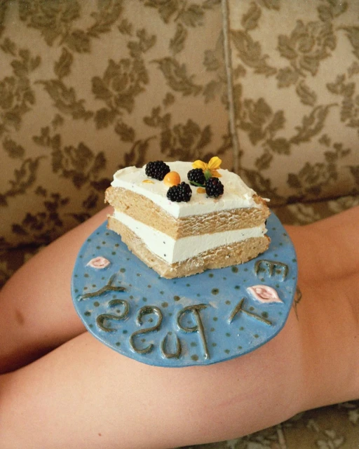 Overhead shot of a layered cake topped with blackberries and a yellow flower on a blue ceramic plate resting on a person's lap.