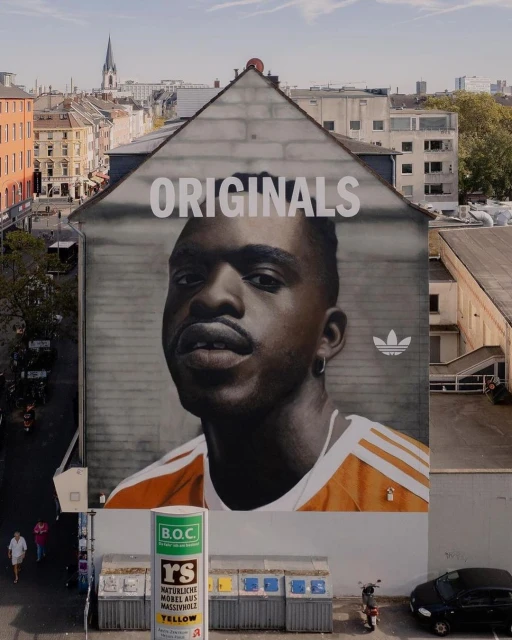 Urban art displays a mural of a man's face on a building with 'ORIGINALS' text, set against a backdrop of buildings in a city.
