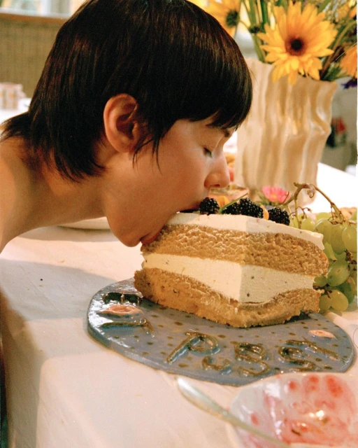 A person eats a large piece of layer cake with blackberries on a blue plate with yellow flowers in the background in an indoor setting.