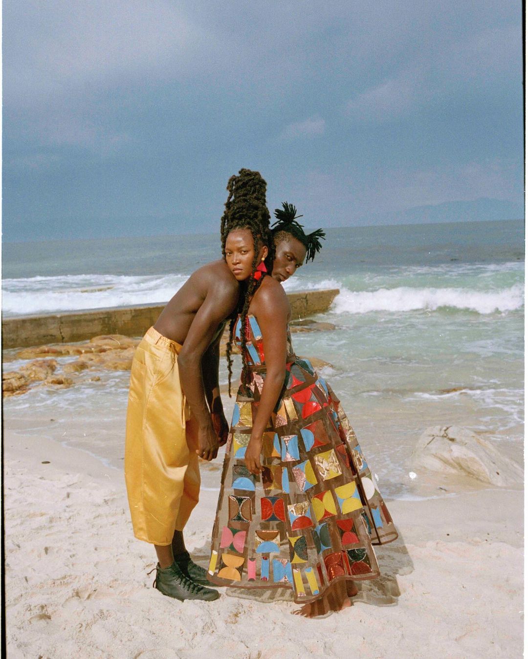 Two people stand close on a sandy beach with ocean backdrop, the person on the right wears a colorful dress. Eye-level angle.