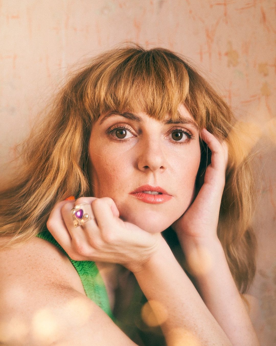 Close-up portrait of a woman with brown hair and a green top, resting her face on her hands, against a textured backdrop with bokeh.