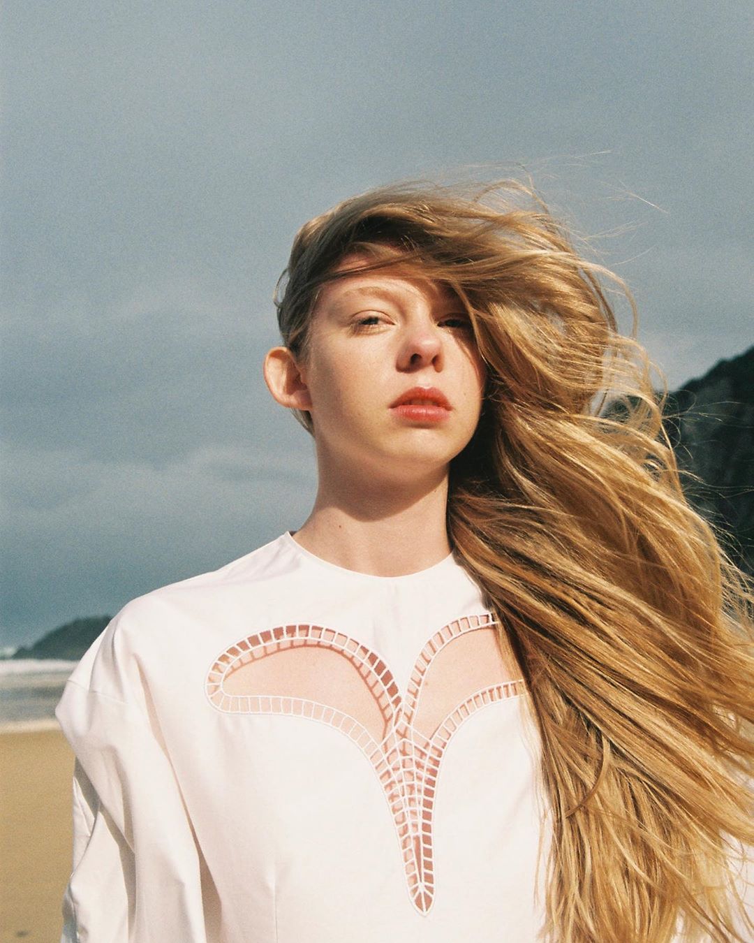 Portrait of a young woman with long blonde hair blowing in the wind, wearing a white top with cut-out details on a beach.