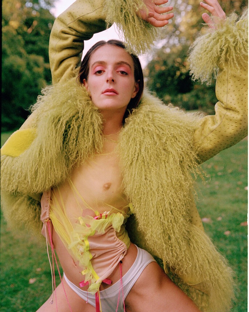 A fair-skinned adult female poses outdoors in an open chartreuse fur coat, light pink eyeshadow, and a revealing light peach-colored outfit.