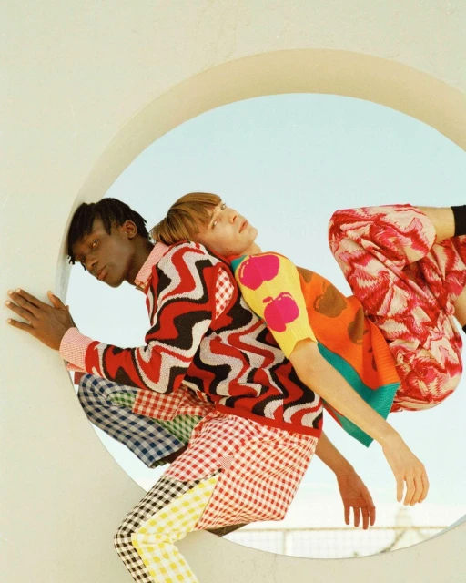 Two fair-skinned and dark-skinned adult fashion models in colorful patterned knitwear pose in front of a circular white structure, outdoors.