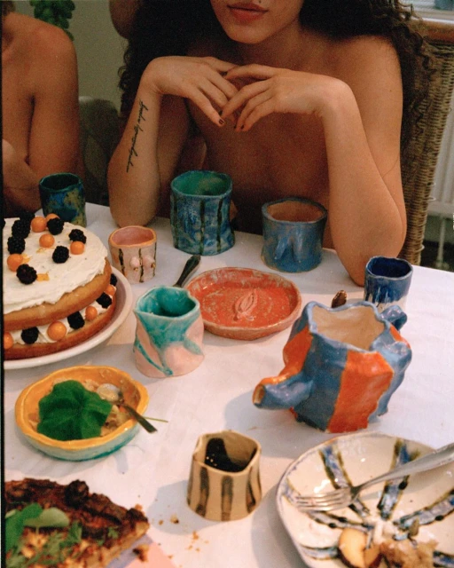 Close-up shot of a table filled with an assortment of colorful handmade ceramic tableware, accompanied by various food items and two people.