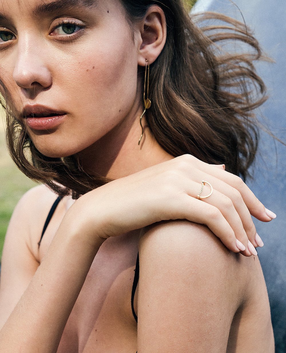 Close-up of a light-skinned young adult woman with brunette hair wearing gold earrings and a ring with an outdoor background.
