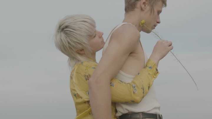 Woman embracing man in outdoor setting. Young couple with short blonde hair, wearing casual clothes. Man holding a flower.