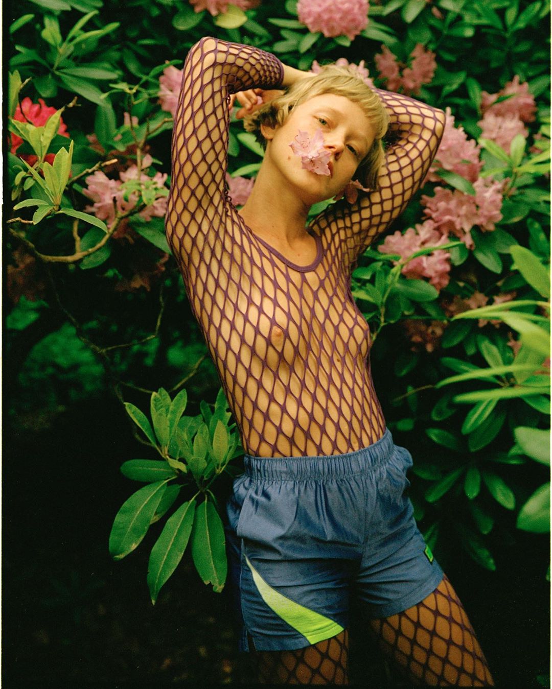 Blonde woman in fishnet top and blue shorts poses among pink rhododendron flowers in a lush green garden. Fashion, portrait, beauty.