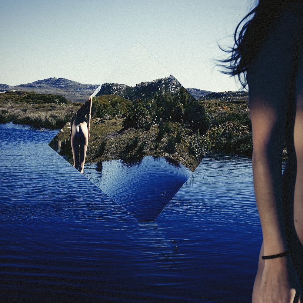 Woman stands near a lake and mountain range. A mirror reflects the landscape, creating a visually striking and surreal outdoor scene.