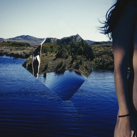 Woman stands near a lake and mountain range. A mirror reflects the landscape, creating a visually striking and surreal outdoor scene.