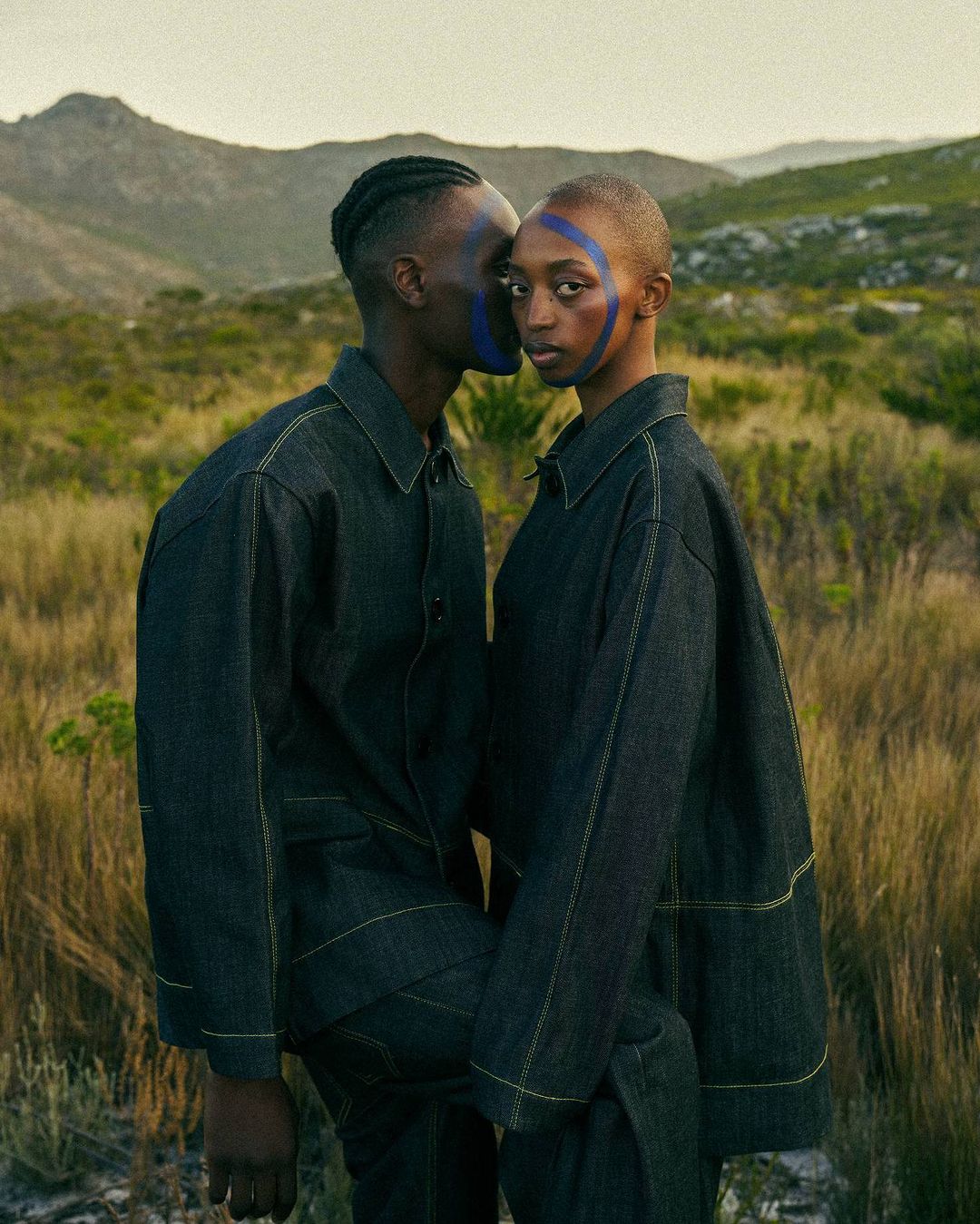 A stylish couple in matching dark denim outfits pose in a grassy field against a mountainous backdrop, showcasing fashion and scenic beauty.