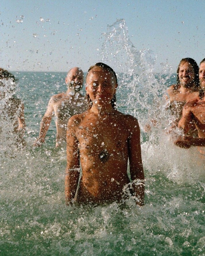 Group of people in the ocean splashing water, including one woman in the center and men with beards, under clear sky and bright lighting.