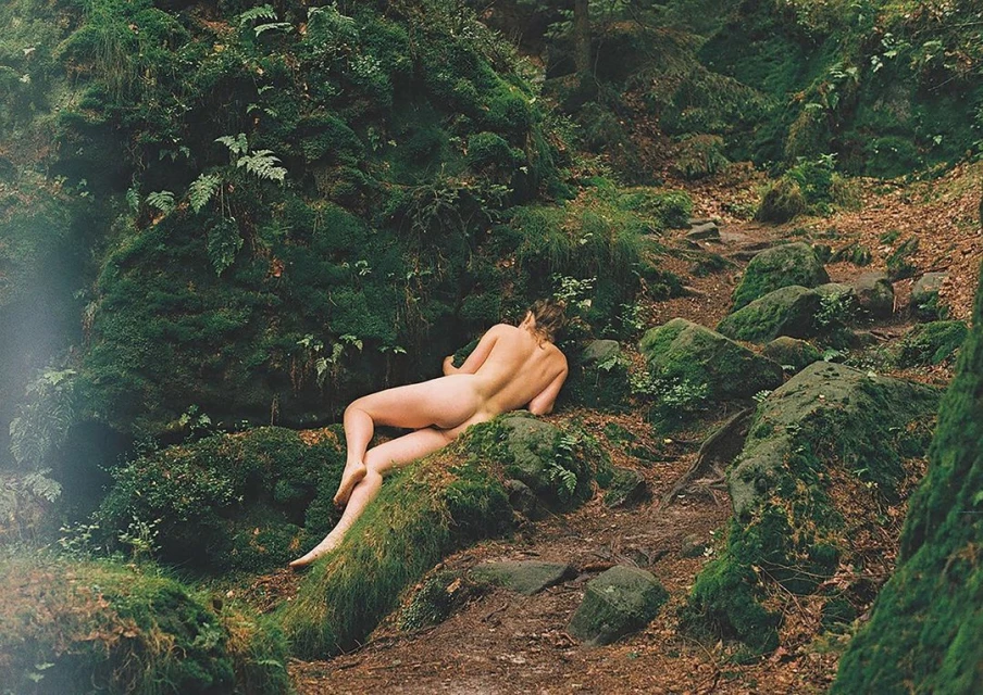 Back view of a nude woman lying on mossy rocks in a forest. Green and brown tones dominate the natural scene, highlighting nature.