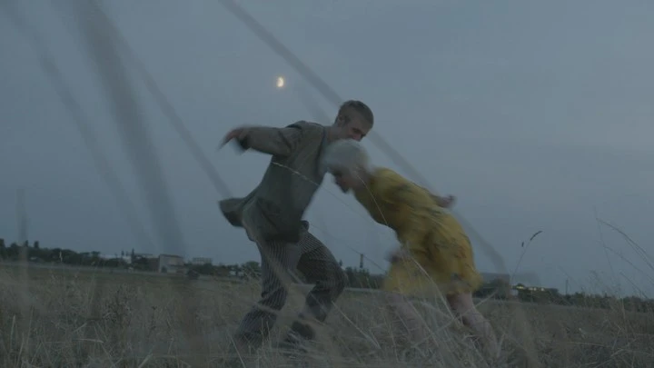 Two people interact in a field at dusk, a man in an olive jacket and striped pants and a woman in a yellow dress.