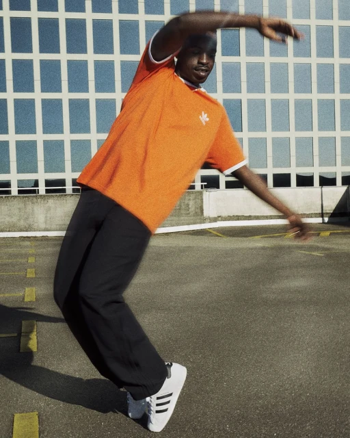 Athletic Black male dancing in orange t-shirt and black pants against a modern building and gray asphalt. Action shot.