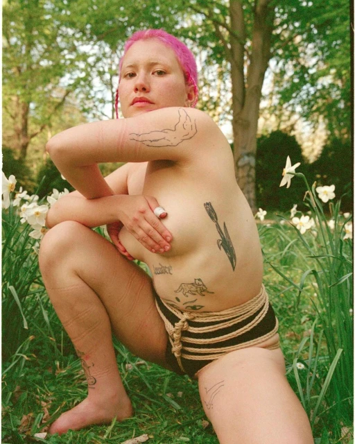 Portrait of a bare-chested person with pink hair squatting in a field of daffodils, showcasing body positivity and natural outdoor setting.