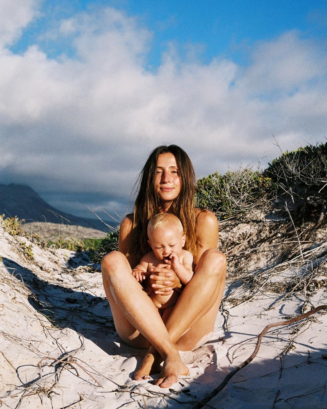 A woman sits outdoors on sandy dunes cradling a baby in her lap, with a backdrop of mountains and a bright sky filled with white clouds.