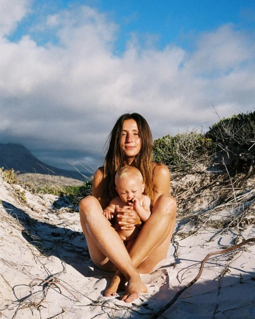 A woman sits outdoors on sandy dunes cradling a baby in her lap, with a backdrop of mountains and a bright sky filled with white clouds.