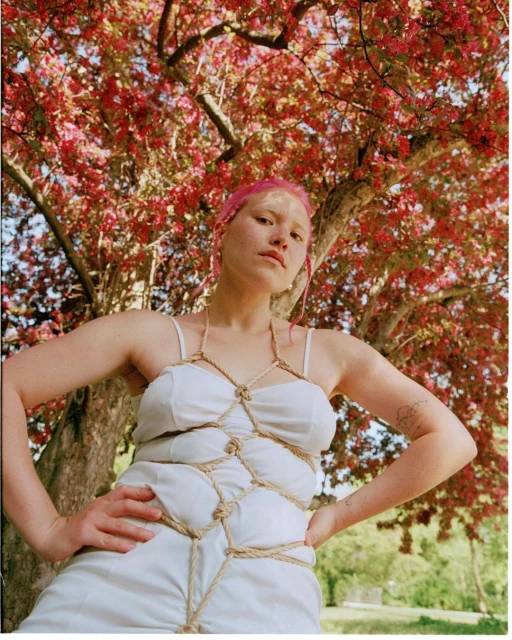 Woman with pink hair in white dress with knotted rope straps poses beneath tree with red blossoms in outdoor, eye-level portrait.