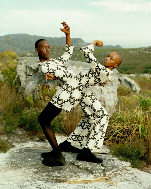 Two people in patterned outfits posing on a rocky surface with grassy vegetation and mountains in the background in a fashion editorial.