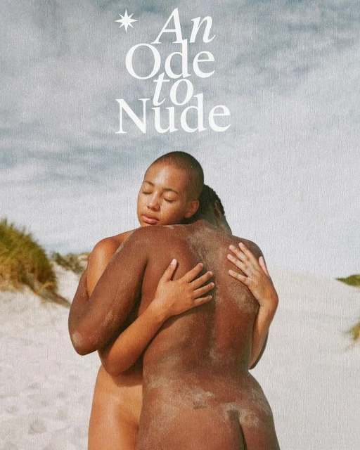 Two figures embrace on a sandy beach under a cloudy sky. One has a shaved head, the other has braided hair. The scene exudes a sense of connection.