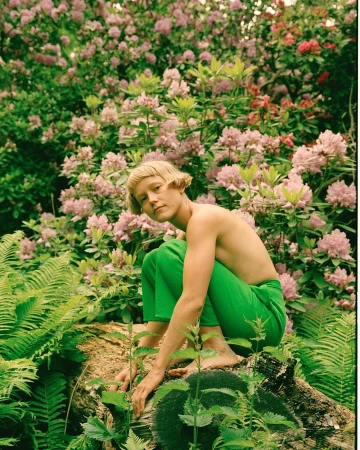 Young adult poses on a log in a green field of ferns and leafy plants, with an abundant backdrop of pink rhododendron blossoms.