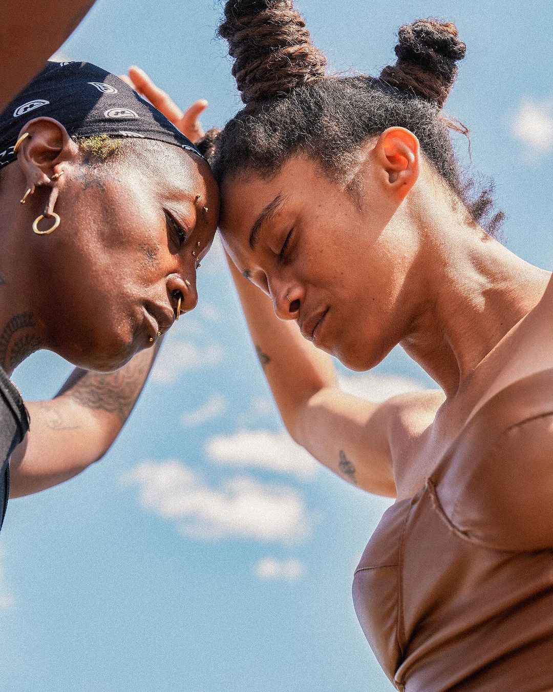 Two adult females face each other outdoors under a blue sky with light clouds, wearing neutral-colored clothing and head coverings in an eye-level shot.