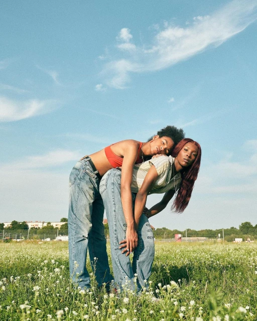Two adult women pose in a sunlit field with blue sky. One woman leans on the other, both wear casual tops and jeans in a meadow.