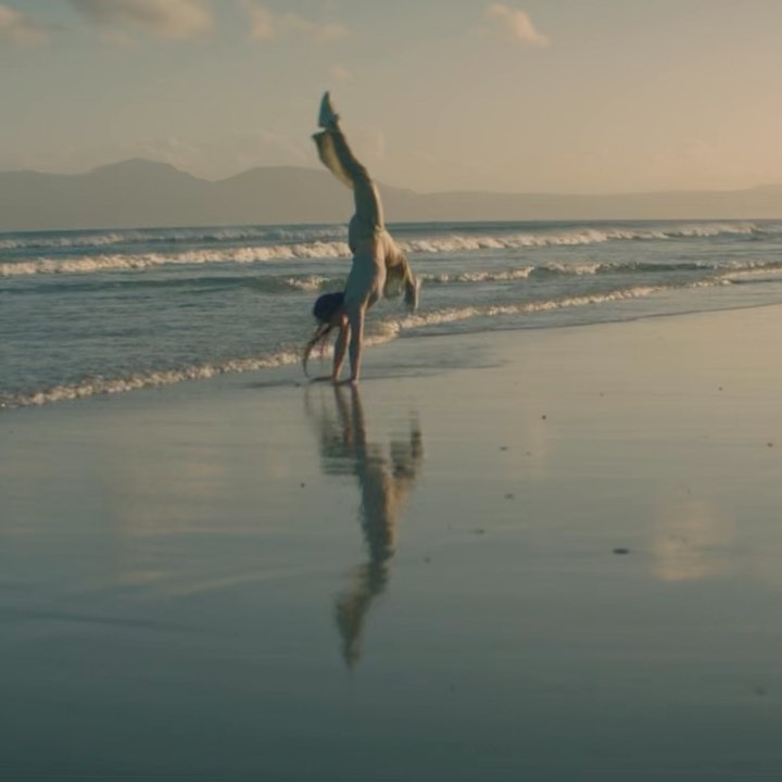 Woman does a handstand on a sandy beach near the ocean and mountains in the background during golden hour.