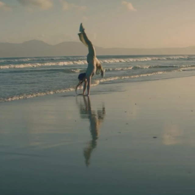 Woman does a handstand on a sandy beach near the ocean and mountains in the background during golden hour.