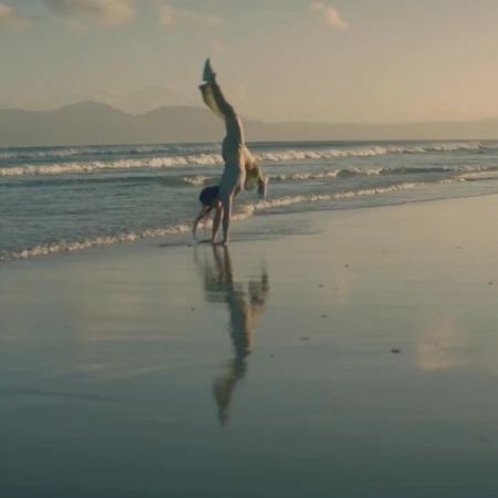 Woman does a handstand on a sandy beach near the ocean and mountains in the background during golden hour.
