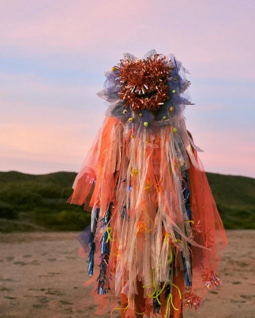 Person in elaborate costume made of orange, white, and blue tulle, tinsel, and other materials, standing on a beach with dunes.