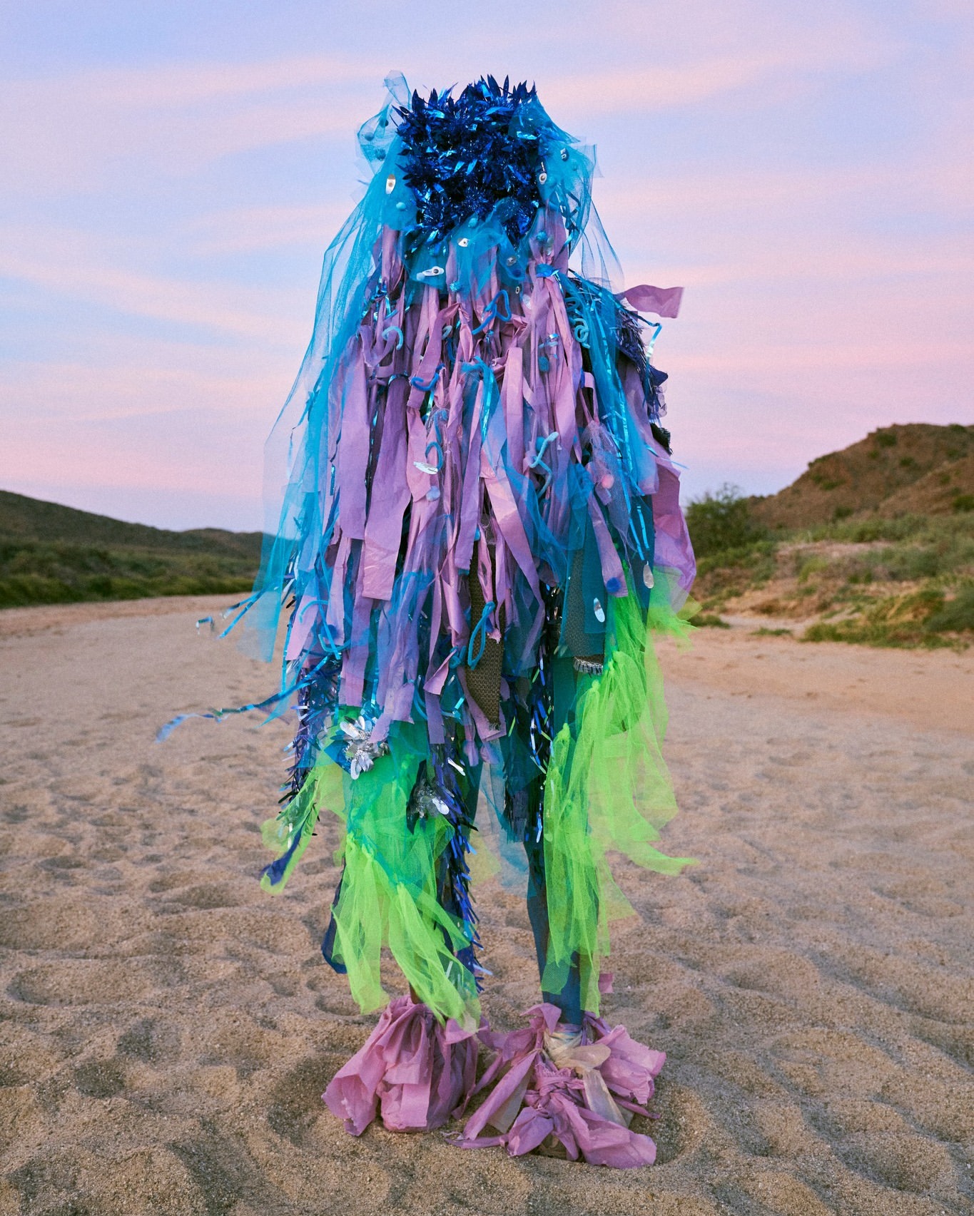 Person in a colorful costume made of blue, purple, and green materials standing on a sandy beach under a pink and blue sky.