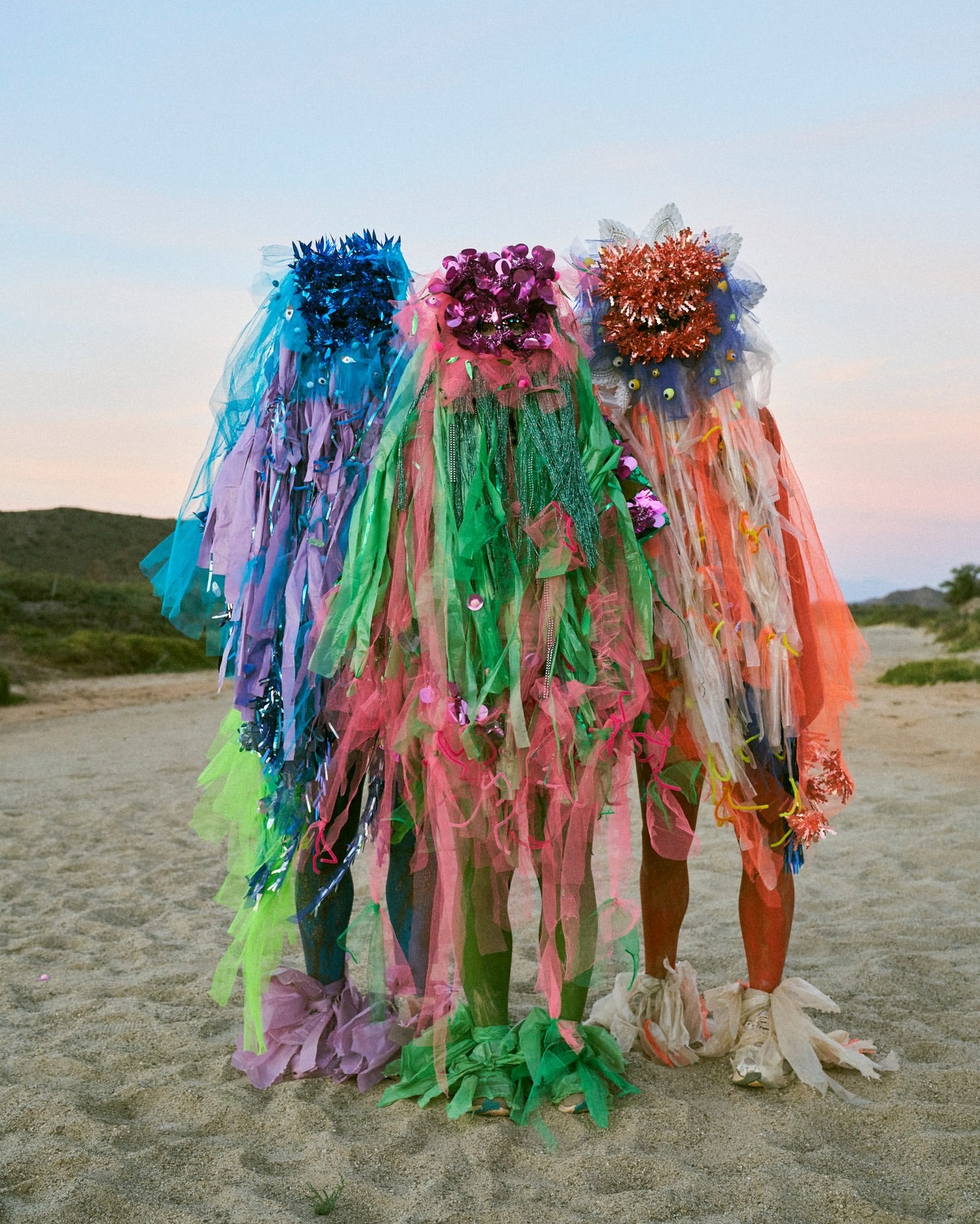 Three figures adorned in colorful, sheer fabric stand on a sandy beach under a pale sky at dusk, presenting an artistic fashion statement.