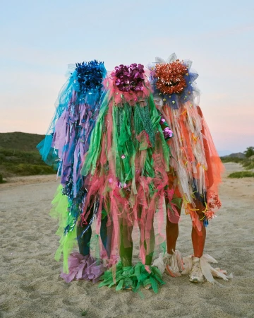 Three figures adorned in colorful, sheer fabric stand on a sandy beach under a pale sky at dusk, presenting an artistic fashion statement.