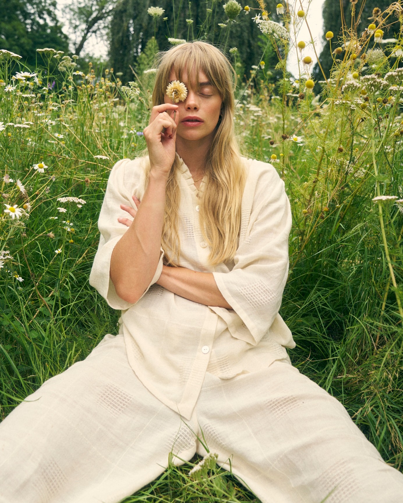 Blond woman in a white outfit poses with closed eyes in a field of wildflowers, holding a daisy in front of her face on a sunny day.