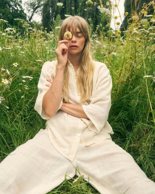 Blond woman in a white outfit poses with closed eyes in a field of wildflowers, holding a daisy in front of her face on a sunny day.