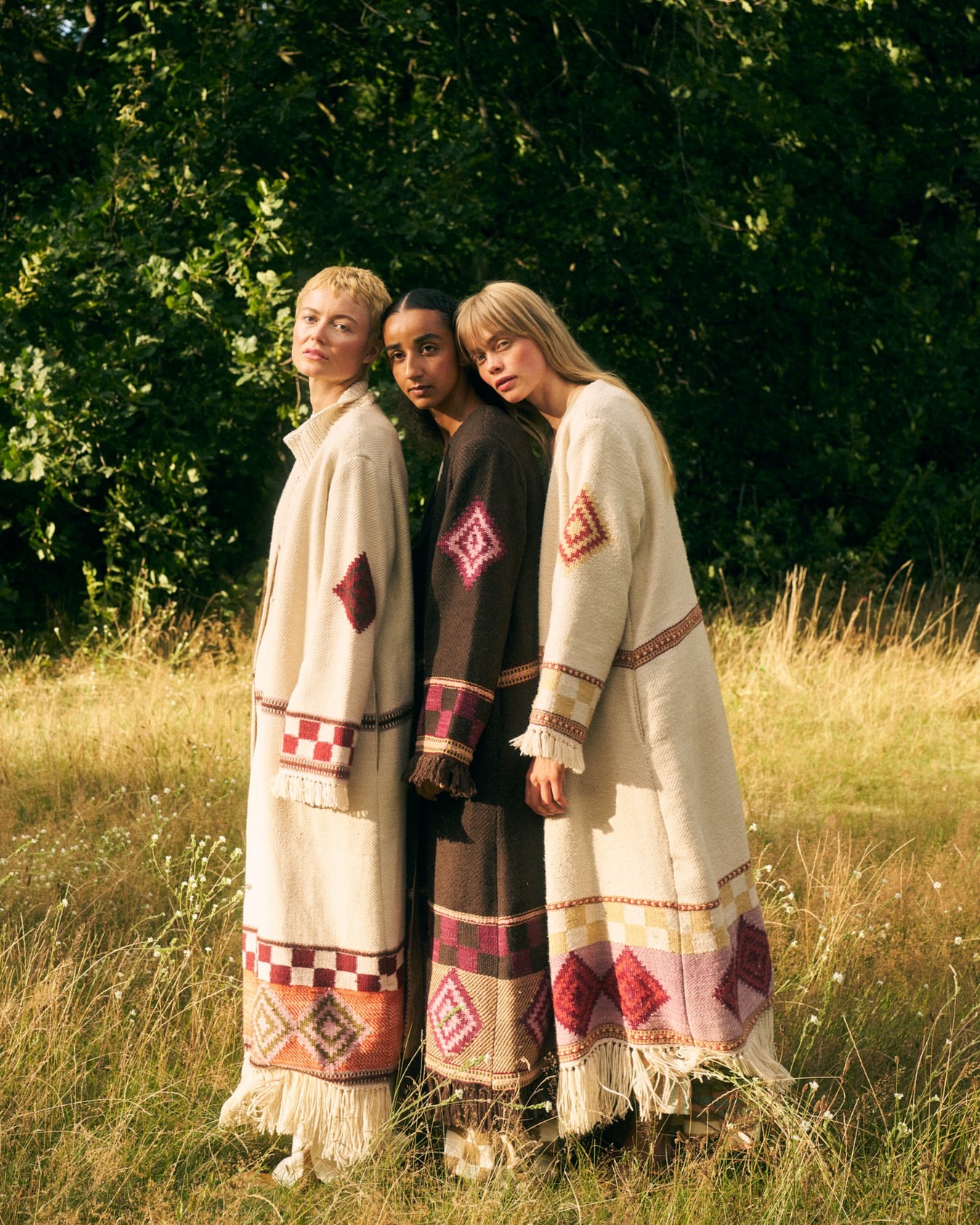 Three female models standing together in a grassy field wearing long patterned coats by photographer Caroline Mackintosh.