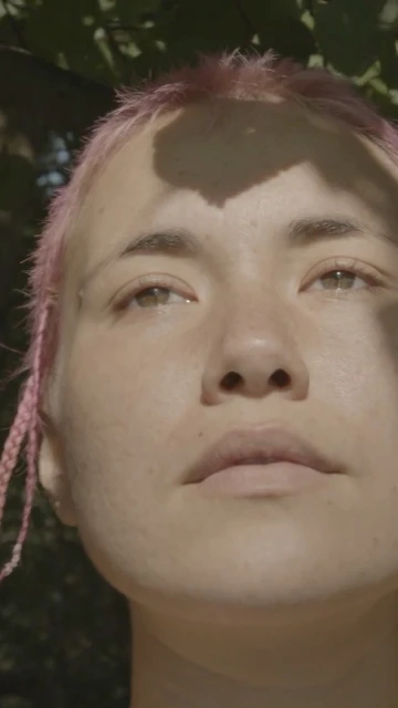 Close-up of a young adult woman with light skin and pink braids looking upwards in an outdoor setting with green foliage.