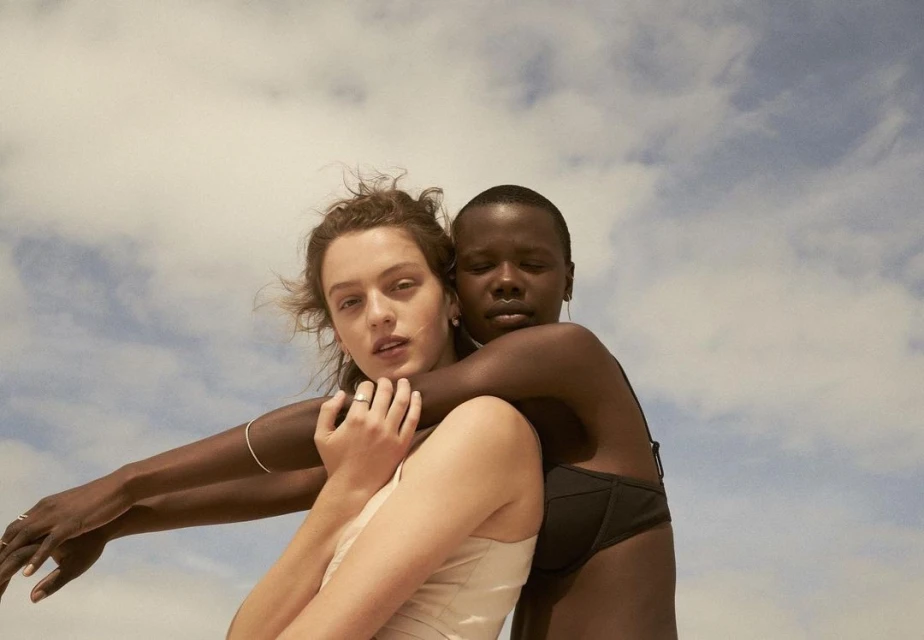 Two women of different ethnicities embrace in an outdoor setting against a bright, cloudy sky. Fashion, friendship, and diversity.